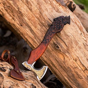 May include: A handcrafted axe with a wooden handle and a decorative wolf head carving. The axe head is silver and gold, and a leather sheath is included. The axe is displayed on a wooden log.
