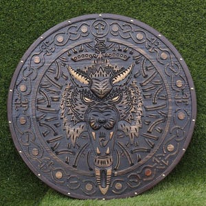 Puede incluir: Un escudo redondo de madera con un diseño tallado de cabeza de lobo. El escudo es de color marrón oscuro con intrincados tallados alrededor de la cabeza del lobo. Los ojos del lobo brillan y el escudo tiene una correa de cuero.