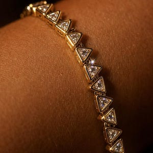 May include: A gold tennis bracelet featuring a series of connected triangular links, each set with a clear, faceted gemstone. The metal has a polished finish, and the stones are arranged in a continuous line, reflecting light with a bright sparkle.