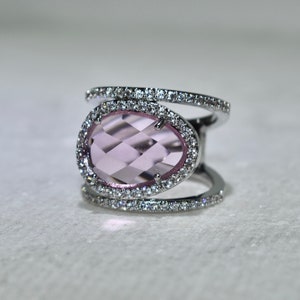 May include: A silver ring with a large, oval-shaped, light pink gemstone surrounded by a row of small, clear gemstones. The ring has a double band design with a row of clear gemstones on each band.