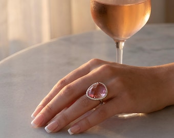 May include: A large, faceted, translucent pink gemstone ring set in a metallic band surrounded by a halo of small, clear, sparkling stones. The ring features an irregular, organic shape and is worn on a finger next to a glass of rose wine.
