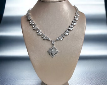 May include: A silver necklace with a diamond-like pendant and accents, displayed on a beige jewelry bust. The necklace features a floral design with a square pendant at the center. The background is a gradient of silver and white.
