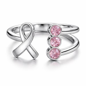 May include: A silver-colored ring featuring a ribbon design and three pink gemstones. The ring has an open design, with the ribbon on one side and the gemstones on the other. The gemstones are round and set in a bezel setting.