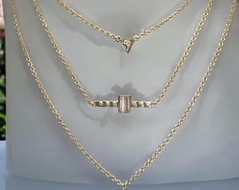 May include: A gold chain necklace with three pendants. The top pendant is a small triangle with a clear stone. The middle pendant is a rectangular bar with a clear stone and small gold accents. The bottom pendant is a larger triangle with a clear stone.
