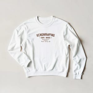 May include: A white sweatshirt with a crew neck and the brown text "KENGNAMPING - EST. 2023 - Never Giving Up". The long-sleeved top has a ribbed neckline, cuffs, and hem. The design is centered on the chest.