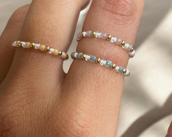 Könnte beinhalten: Vier Perlenringe in verschiedenen Farben werden auf einem Finger gezeigt. Die Ringe sind mit kleinen, farbigen Perlen in Gold-, Weiß-, Pfirsich-, Rosa- und Blautönen verziert. Die Ringe wirken zart und scheinen handgefertigt zu sein, was einen minimalistischen Schmuckstil zeigt.