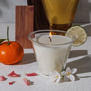 May include: A white candle in a clear glass jar with a black wick burning. The candle is surrounded by pink flower petals, a small orange, and a lime slice.