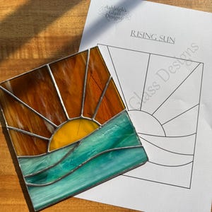 Può includere: Un sole nascente in vetro colorato sopra l'oceano. Il sole è giallo con raggi arancioni. L'oceano è blu turchese con onde. Il vetro è posto su un motivo di carta bianca con le parole "Rising Sun" e "Ashleigh's Glass Designs".