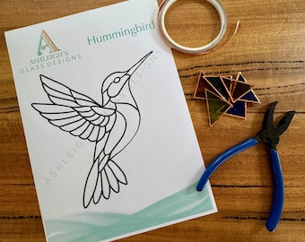 May include: A stained glass craft kit featuring a hummingbird design. The kit includes a printed design, copper foil tape, a small stained glass piece, and blue-handled pliers. The design is from Ashleigh's Glass Designs.