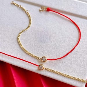 May include: Two delicate gold bracelets with a red string and a small, round, clear gemstone. One bracelet is a gold chain, and the other is a red string with a gold chain accent. The bracelets are on a white surface.