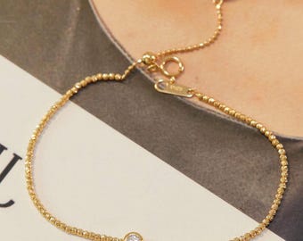 May include: A delicate gold bracelet with a small, round diamond at the center. The bracelet features a beaded chain design and an adjustable clasp. The gold color is bright and shiny, reflecting light. This piece of jewelry is elegant and suitable for everyday wear.