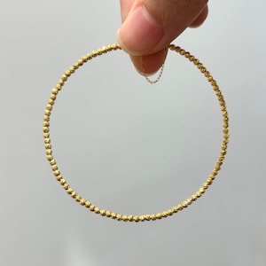 May include: A gold beaded bracelet with a delicate chain clasp. The bracelet is made of small, evenly spaced beads, creating a textured surface. The bracelet is held up by a hand against a plain background.