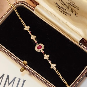 Op de afbeelding: Een goudkleurige armband met een ovale robijn in het midden en kleine diamanten. De armband wordt gepresenteerd in een vintage sieradendoosje met een zwarte fluwelen binnenkant. De ketting is delicaat.