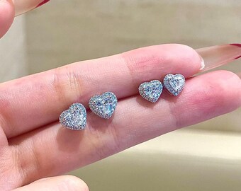 May include: Close-up of three pairs of heart-shaped stud earrings. Each earring features a central, clear gemstone surrounded by a halo of smaller, clear stones. The earrings are displayed on a light-toned background.