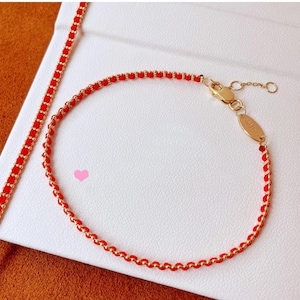May include: A red and gold bracelet with a delicate chain design. The bracelet features small red beads woven with gold-coloured metal. The clasp and a small tag are also gold. The bracelet is displayed on a white surface.