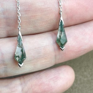 Może przedstawiać: Para srebrnych kolczyków wiszących z zielonymi kroplami agatu mchowego. Kropelki są fasetowane i mają kształt łzy.