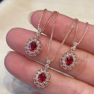 Peut inclure: Trois colliers en or rose avec des pierres précieuses ovales rouge foncé, entourées de petites pierres claires. Les pendentifs sont suspendus à de fines chaînes. Les bijoux sont présentés sur un fond neutre.