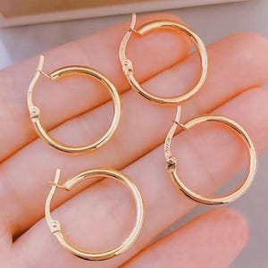 May include: Four gold hoop earrings. The earrings are round and have a simple, classic design. The hoops are a warm, shiny gold color. The earrings are shown on a light-colored background.
