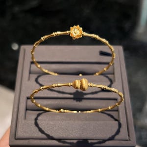 May include: Two gold-colored bangles. The top bangle features a flower design, while the bottom bangle has a double-sphere center. Both are crafted with a bamboo-like texture, presented on a gray display.