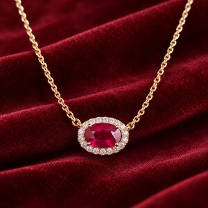 May include: A gold chain necklace featuring an oval-shaped red gemstone pendant surrounded by a halo of small, clear, sparkling stones. The pendant is centered on a delicate gold link chain that connects to both sides of the oval setting.