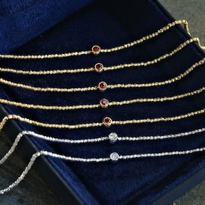 May include: Several delicate necklaces presented in a dark blue box. The necklaces have thin, textured chains. Some are gold with red gemstones, others silver with clear gemstones, demonstrating a minimalist jewellery design.