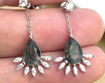 May include: A pair of silver dangle earrings featuring green moss agate pear-shaped stones and a halo of clear cubic zirconia stones.