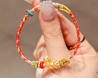 May include: A braided red and gold bracelet with gold accents and a small gold horse charm. The bracelet features a mix of red, gold, and white threads, with additional decorative beads. The bracelet is held by a hand.