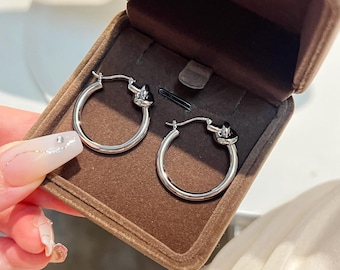 May include: A pair of silver hoop earrings with a knot design, displayed in a brown jewelry box. The earrings have a polished finish and a classic, elegant style. The box is open, revealing the earrings against a soft, velvety interior.