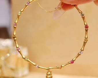 May include: A gold-colored bracelet with a bamboo design, interspersed with small, colorful beads. The bracelet features a small charm of a cat. The bracelet is held up by a person's hand.
