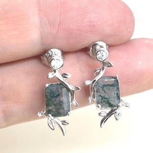 May include: Silver earrings with a green moss agate stone and a small clear crystal. The earrings feature a delicate branch design with leaves.