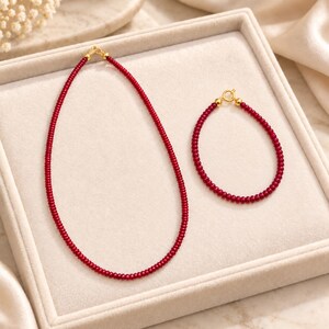Könnte beinhalten: Ein Schmuckset bestehend aus einer Halskette und einem Armband, beide gefertigt aus kleinen, gleichmäßigen, dunkelroten Perlen. Jedes Stück verfügt über einen goldfarbenen Metallverschluss und kleine goldfarbene Akzentperlen an den Enden.