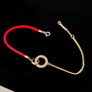 Peut inclure: Un bracelet doré avec un cordon rouge et une fine chaîne. Le bracelet présente deux cercles entrelacés, l'un avec un motif de diamants et l'autre avec du texte gravé. Le fond est noir.