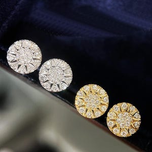 Puede incluir: Cuatro pendientes de botón redondos. Dos son plateados y dos dorados. Cada pendiente presenta un grupo central de pequeñas piedras transparentes rodeadas por un patrón de estrella de piedras transparentes más grandes, creando un efecto brillante. Los pendientes se muestran sobre una superficie azul oscuro.