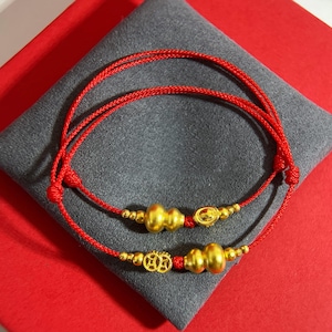 Könnte beinhalten: Zwei rote Schnurarmbänder mit goldfarbenen Anhängern. Die Anhänger umfassen Kürbisformen und dekorative kreisförmige Elemente. Die Armbänder werden auf einem grauen Kissen auf einer roten Oberfläche präsentiert.