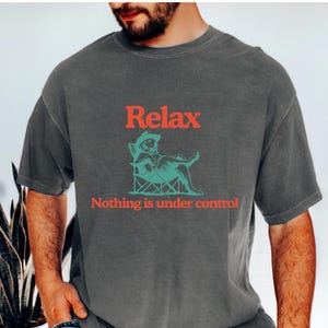 May include: Dark gray t-shirt with the word "Relax" in red above a turquoise illustration of a figure reclining in a chair. Below the illustration, the text reads "Nothing is under control."