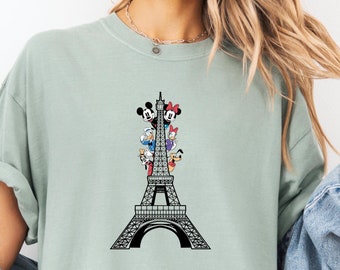 May include: A light green t-shirt featuring a black and white graphic of Mickey Mouse, Minnie Mouse, Donald Duck, Goofy, and Pluto standing on the Eiffel Tower.