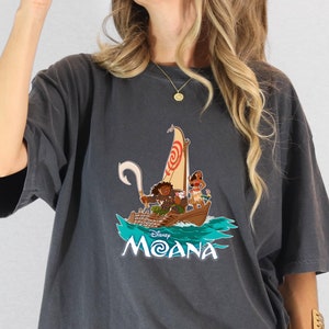 May include: A dark gray t-shirt with a graphic print of the Disney characters Moana and Maui sailing on a boat. The text "Disney Moana" is printed below the image.