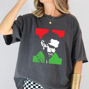 May include: Dark grey t-shirt featuring a graphic of a man with glasses, a green jacket, and red and green shapes. The design is a stylized portrait. The shirt has short sleeves and a relaxed fit.