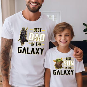 Pu&ograve; includere: Magliette bianche con grafica abbinata. Una maglietta dice "BEST DAD IN THE GALAXY" con una grafica di Mandaloriano. L'altra dice "BEST SON IN THE GALAXY" con una grafica di Grogu. Le magliette sono indossate da un padre e un figlio.