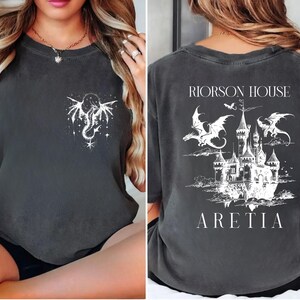May include: Dark gray t-shirt with a white dragon and star design on the front. The back features a castle illustration with dragons, the text "RIORSON HOUSE" above, and "ARETIA" below. A fantasy-themed graphic tee.