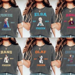 May include: Six gray t-shirts with different Frozen characters printed on them. Each shirt has the text "EST 2013 FROZEN" printed below the character. The characters are Princess, Elsa, Anna, Hans, Olaf, and Kristoff.