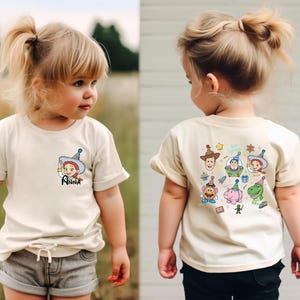Puede incluir: Camiseta color crema con una imagen de dibujos animados de un personaje con el nombre "Anna" en la parte delantera. La parte posterior de la camiseta muestra varios personajes de dibujos animados. La camiseta la lleva un ni&ntilde;o peque&ntilde;o.
