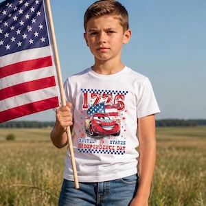 A young boy wearing a white t-shirt with a graphic of a red car and the text "1775 United States Independence Day" is standing in a field, holding an American flag.