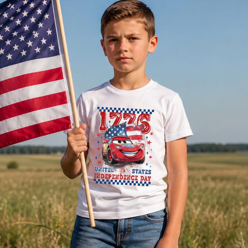 A young boy wearing a white t-shirt with a graphic of a red car and the text "1775 United States Independence Day" is standing in a field, holding an American flag.