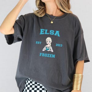 May include: A charcoal gray t-shirt with a blue graphic of Elsa from Frozen. The graphic includes the text "Elsa Est 2013 Frozen".