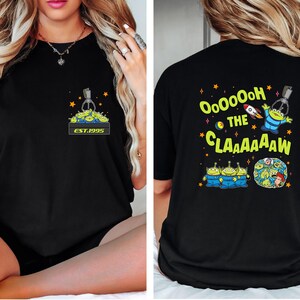 May include: Black t-shirt featuring a graphic of the claw machine with green aliens and the text "EST.1995" on the front. The back of the shirt has the text "Ooooooh the Claaaaaw" with alien characters and a rocket ship.