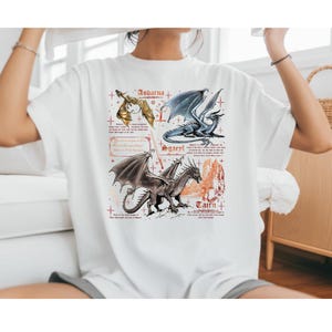 Peut inclure: T-shirt blanc avec trois illustrations de dragons nomm&eacute;s "Andarna", "Sgaerl" et "Tairn". Les dragons sont repr&eacute;sent&eacute;s en or, bleu et gris, avec des ailes et des &eacute;cailles d&eacute;taill&eacute;es. Du texte est inclus sous chaque dragon.