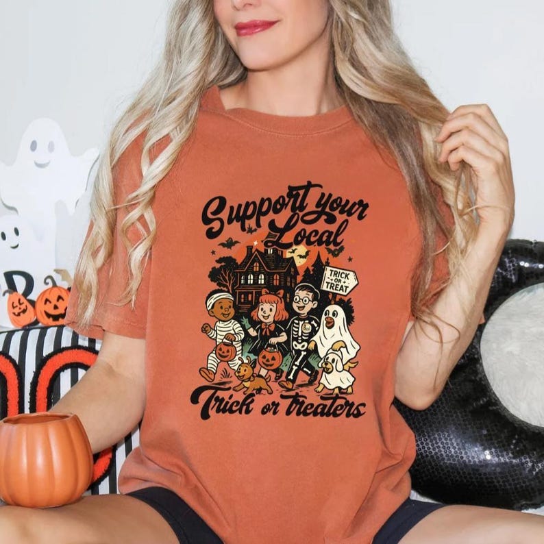A woman wearing a t-shirt with a Halloween-themed graphic and text, sitting in front of Halloween decorations.