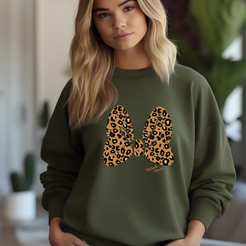 May include: An olive green sweatshirt featuring a large bow design with a leopard print pattern. The bow is centered on the front of the sweatshirt. The text "Minnie Mouse" is printed in a cursive font below the bow.