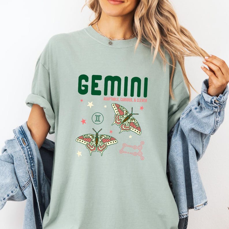 Może przedstawiać: Jasnozielona koszulka z napisem "GEMINI" w ciemnozielonych, blokowych literach. Poniżej słowa "ADAPTABLE, CURIOUS, & CLEVER". Koszulka przedstawia dwie ilustracje motyli i wz&oacute;r konstelacji.